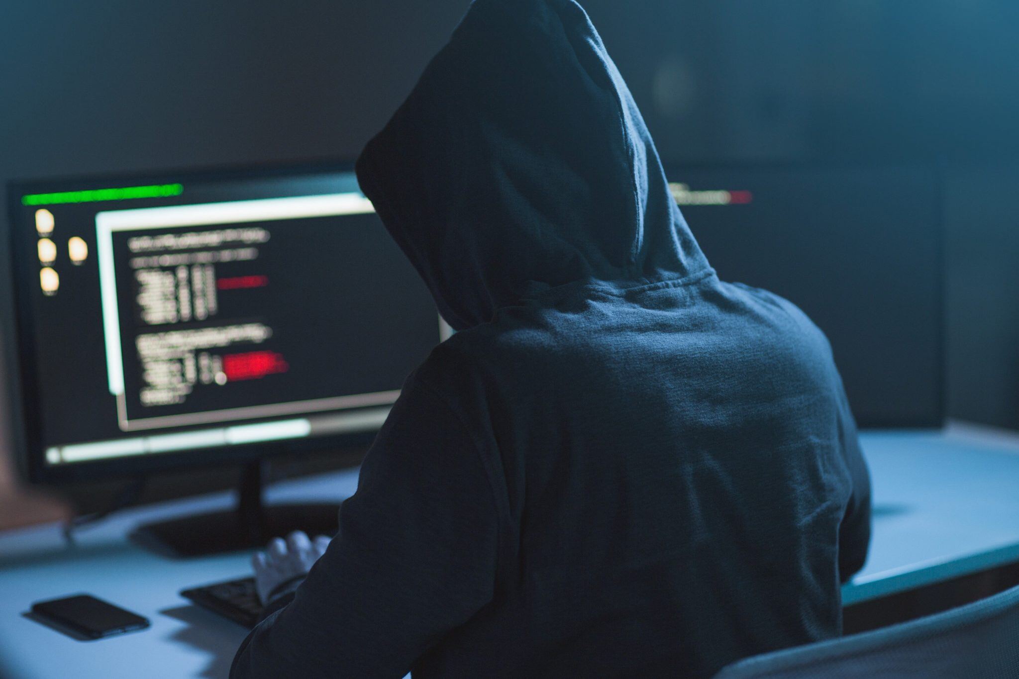 A hooded figure silhouetted at a monitor displaying lines of code